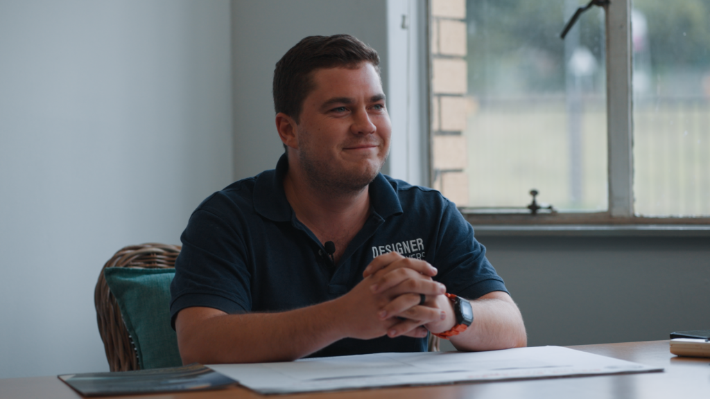 designer pool covers A man wearing a "designer" polo shirt sits at a table, smiling slightly, with his hands clasped in front of him and papers beneath them. there is a window in the background.